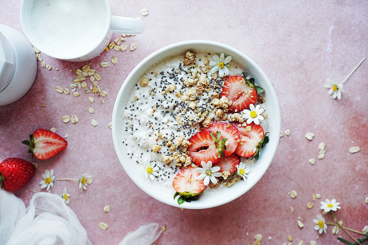 A bowl with Oatmeal Greek Yogurt topped with strawberries set on a pink background with a coffee mug on the side. 