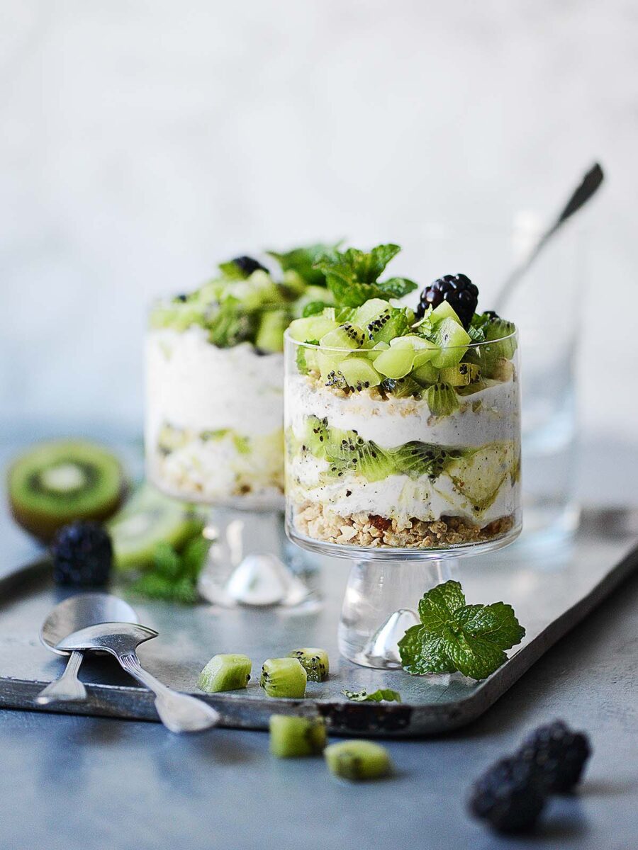 Two cute glasses with Granola & Yogurt parfait topped with fresh kiwi placed on a metal tray.